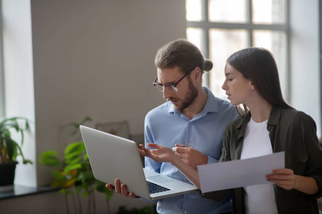 male and female coworkers standing in office discussing disaster recovery plan on laptop