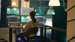 Cybercriminal sitting in front of multiple monitors hacking businesses with poor cybersecurity